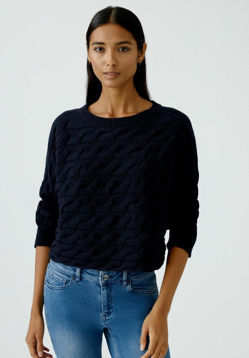 Marineblauer Pullover mit strukturiertem Kabelstrickdesign, Rundhalsausschnitt und dreiviertellangen Ärmeln, kombiniert mit hellblauen Jeans.