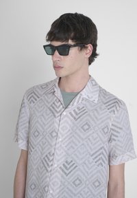 Camicetta a maniche corte di un leggero rosa pallido con un motivo geometrico in tonalità di grigio, abbinata a occhiali da sole neri angolari. Canottiera grigia semplice visibile.