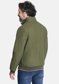 Chaqueta bomber verde oliva con un cuello alto, que presenta una textura suave y puños acanalados. Llevada sobre unos jeans azules, vista por la parte de atrás.