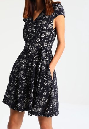 Femme portant une robe noire à fleurs à manches courtes avec un col en V et une taille nouée, debout avec une main dans sa poche.