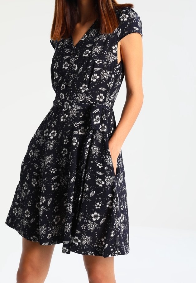 Femme portant une robe noire à fleurs à manches courtes avec un col en V et une taille nouée, debout avec une main dans sa poche.
