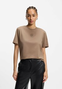 T-shirt in cotone marrone con maniche corte, collo a giro e un piccolo logo ricamato sul petto. Abbinato a pantaloni in pelle nera lucida.