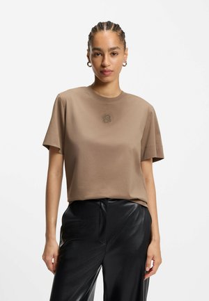 T-shirt in cotone marrone con maniche corte, collo a giro e un piccolo logo ricamato sul petto. Abbinato a pantaloni in pelle nera lucida.