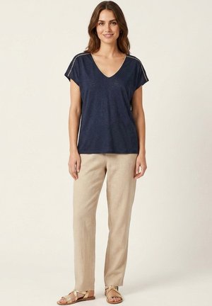 Femme debout, portant un haut bleu marine à col en V et manches courtes, un pantalon beige droit et des sandales beiges à brides, sur un fond clair uni.