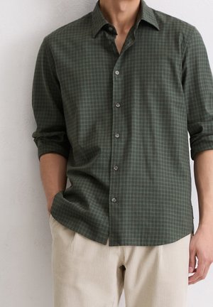 Mężczyzna ubrany w ciemnozieloną kraciastą koszulę z podwiniętymi rękawami i beżowe spodnie sztruksowe, stojący przy gładkiej ścianie.