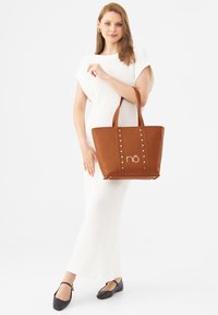 Braune vegane Ledertasche mit goldenen Nieten an den Seiten, einem Logo auf der Vorderseite und zwei stabilen Griffen. Glatte Textur und strukturierte Form.