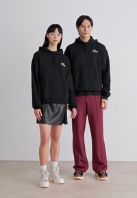 Sudaderas con capucha negras y bolsillos frontales, usadas sobre prendas oscuras. Modelo 1 con una falda de cuero y deportivas; Modelo 2 con pantalones burdeos y zapatos.