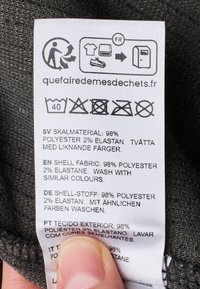 Pflegeetikett auf Stoff, weiß mit schwarzem Text, der die Zusammensetzung angibt: 98% Polyester, 2% Elastan; enthält Waschhinweise und Symbole.