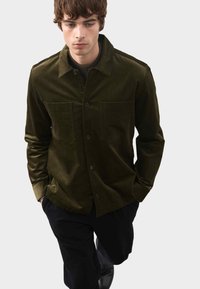 Tkanina kordowa w kolorze oliwkowym, kurtka koszulowa z dwoma kieszeniami na piersi, zapięcie na guziki i klasyczny kołnierzyk. Długie rękawy i miękka tekstura.