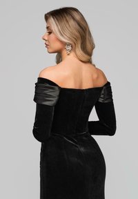 Czarna sukienka z aksamitu z odkrytymi ramionami, satynowymi rękawami, dopasowanym gorsetem oraz długą, gładką spódnicą. Kolczyki zdobione są detalicznym haftem.