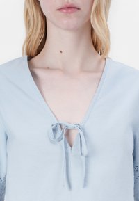 Γαλάζιο μπλουζάκι κοντό μανίκι με V λαιμόκοψη και λεπτομέρεια με κορδόνι μπροστά. Διαθέτει ανοιχτές λεπτομέρειες στα μανίκια. Μαλακή υφή υφάσματος.