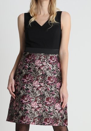 Femme portant une robe noire sans manches avec une jupe florale texturée dans des tons de rose, bordeaux et argenté, debout devant un fond uni.