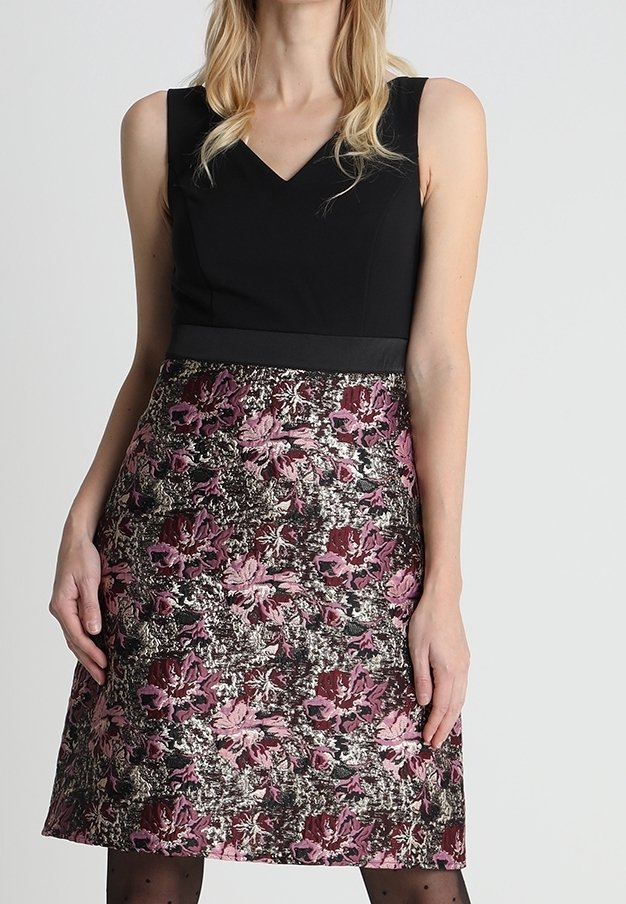 Femme portant une robe noire sans manches avec une jupe florale texturée dans des tons de rose, bordeaux et argenté, debout devant un fond uni.