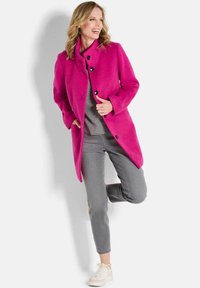 Cappotto di lana fucsia con collare alto, design monopetto, grandi bottoni neri e due tasche laterali. Indossato sopra un top grigio e abbinato a pantaloni grigi.
