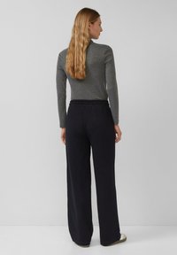 Kobieta z długimi blond włosami, ubrana w szary długi rękaw, luźne czarne spodnie, stoi przodem do gładkiej jasnoszarej ściany.