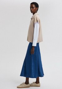 Beige mouwloze top over een wit shirt met lange mouwen, gecombineerd met een blauwe denimrok en beige sneakers, met een uitlopende zoom en stikseldetails.