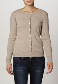 Beige stickad kofta med långa ärmar, rund halsringning och knäppning fram. Har ribbade manschetter och fåll. Mjuk textur.