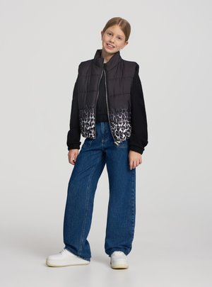 Ragazza giovane che sorride, indossa un gilet imbottito nero con stampa leopardata, una maglia nera a maniche lunghe, jeans larghi blu e scarpe da ginnastica bianche su uno sfondo uniforme.