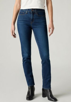 Kvinde iført slim fit blå jeans, hvid top og sorte ankelstøvler med hæl, står foran en ensfarvet baggrund.