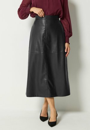 Vrouw draagt een zwarte leren midi-rok met hoge taille en naden, gecombineerd met zwarte puntige hakken en een bordeauxrode blouse met lange mouwen.