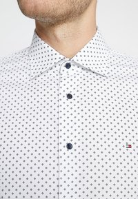 Weißes Hemd mit kleinem Diamantmuster, dunkelblauen Knöpfen und einem Kentkragen. Mit dezentem Logo der Marke an der Seite.