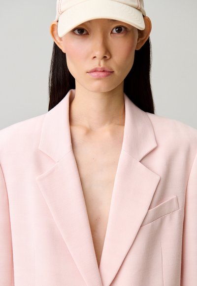 Jeune femme aux longs cheveux bruns portant un blazer rose pâle oversize et une casquette de baseball beige clair sur un fond neutre.