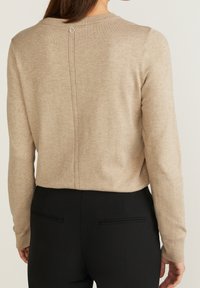Beige Strickpullover mit rundem Halsausschnitt, langen Ärmeln und einer Mittelnaht in der Rückseite, die mit einem kreisförmigen Akzent versehen ist. Kombiniert mit schwarzen Hosen.