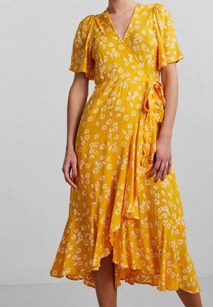 Frau trägt ein gelbes Wickelkleid mit Blumenmuster, kurzen Schmetterlingsärmeln und einem asymmetrischen, gerafften Saum, der in der Taille gebunden ist, vor hellem Hintergrund.