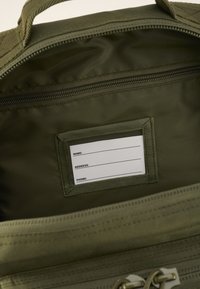 Alyvuogių žalios spalvos kuprinės vidus, kuriame yra užtrauktuko kišenė ir etiketės laikiklis su tuščiomis laukeliais vardui, adresui ir telefono numeriui. Struktūrizuota medžiaga.