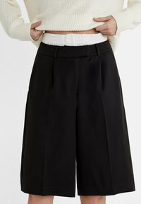 Pantalones cortos negros con pliegues, de corte relajado y cinturilla elástica. Fabricados en material ligero, con una textura suave y un diseño confeccionado.