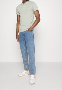 Světle zelené polo tričko s krátkými rukávy, světle modré džíny se širokými nohavicemi a bílé tenisky s oranžov ými detaily. Jemné logo na polo tričku.