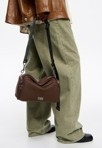 Ruda audinio peties rankinė su banginiu dizainu, užtrauktukais viršuje ir juodu reguliuojamu diržu. Priekyje yra "BIMBA Y LOLA" logotipas.