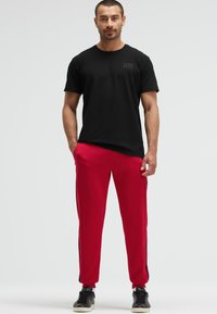 Schwarzes Baumwoll-T-Shirt mit rundem Halsausschnitt, kombiniert mit roten Jogginghosen mit Seitenteilen und schwarzen Akzenten. Getragen mit schwarzen Sneakern.