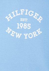 T-shirt di cotone azzurro chiaro con testo bianco: "HILFIGER EST. 1985 NEW YORK" in grassetto, disposto in un layout circolare. Tessuto morbido e design casual.
