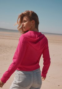 Crop top rose à capuche avec tissu texturé, assorti à un short en jean clair. La scène se déroule sur une plage sous un ciel bleu clair.