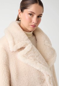 Giovane donna con capelli slicked-back che indossa orecchini d'oro, un dolcevita crema e un morbido cappotto shearling beige con ampi risvolti.