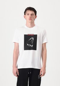 Hvit bomull t-skjorte med et svart kvadratisk grafikk av en munn med tenner, og merkets navn "HUGO" i rødt over.
