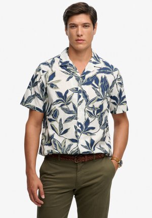 Hombre con camisa blanca de manga corta abotonada con estampado de hojas azules, pantalones verde oliva, cinturón trenzado marrón y reloj de pulsera, de pie con la mano en el bolsillo.