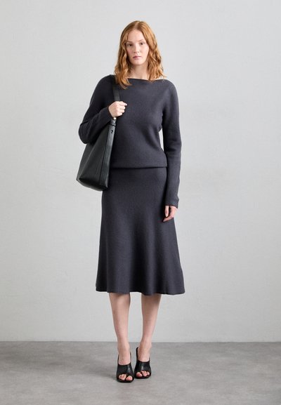 Dunkelgrauer Strickpullover und passender knie-langer Rock, beide mit einer weichen Textur. Das Modell hält eine schwarze Tragetasche und trägt schwarze Sandalen mit offenem Zehenbereich.