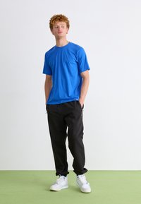 Camiseta de manga corta azul, hecha de tela suave, combinada con joggers negros. Calzado blanco con detalles. Fondo liso y suelo verde.