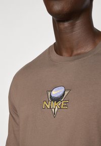 T-shirt en coton marron avec un logo Nike multicolore brodé en forme de triangle et un motif de balle de golf sur la poitrine.