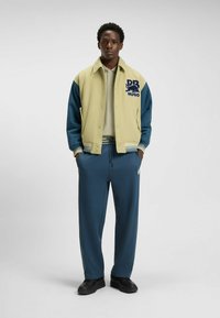 Kremowa i miętowa kurtka typu varsity z haftem, zestawiona z luźnymi miętowymi spodniami i czarnymi butami. Kurtka ma ściągacze przy mankietach i kołnierzu.