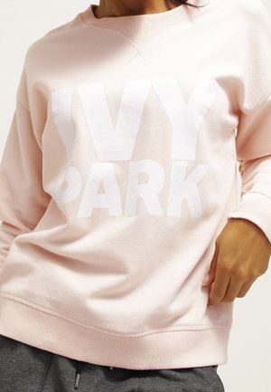 Osoba nosząca jasnoróżową bluzę z dużym białym napisem "IVY PARK", w połączeniu z ciemnoszarymi spodniami, ręka oparta blisko pasa.