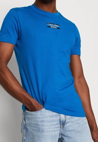 Modré bavlněné tričko s kulatým výstřihem a obdélníkovou nášivkou s logem "Calvin Klein Jeans" v černé a bílé barvě.