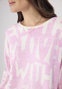 Rosa und cremefarbener Sweatshirt mit großem, abstraktem Buchstabenmuster. Verfügbar mit Rundhalsausschnitt und langen Ärmeln. Sichtbare goldene, mehrlagige Kettenkette.