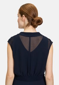 Blouse marine avec des manches courtes en dentelle et un décolleté en V dans le dos ; présente une taille smockée pour un ajustement et un mouvement fluides. Tissu lisse et léger.