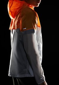 Chaqueta ligera con capucha naranja y cuerpo gris. Tejido texturizado con detalles reflectantes en las mangas. Diseño de media cremallera.