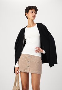 Crna oversized jakna, bijela uska majica s dugim rukavima, bež mini suknja od kordura s gumbima sprijeda i svijetlo bež torba.
