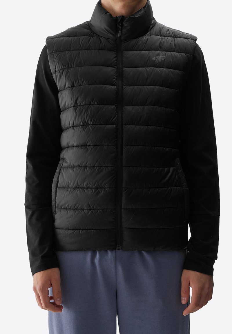 Schwarze gepolsterte Weste mit hohem Kragen und durchgehendem Reißverschluss, mit glatter Textur und integrierten Taschen, die über einem langen schwarzen Oberteil getragen wird.
