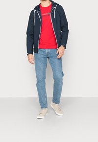 Marineblå zip-up jakke med hætte, hvide snore, båret over en rød skjorte og lyseblå jeans, parret med beige sneakers. Minimalistisk design.